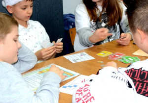 Grupa uczniów gra przy stoliku. Uczniowie trzymają karty do gry. Na stoliku znajdują się karty z rysunkami strzałek oraz plansza do gry.